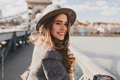 Outdoor photo of romantic european woman with curly hairstyle spending time outdoor, exploring european city. Graceful young lady in gray coat and hat enjoying views on embankment.