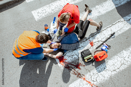 Ambulance worker with man in road vest applying emergency medical care to the injured bleeding person lying on the pedestrian crossing after the accident, view from the above