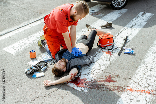 Ambluence worker applying emergency care to the injured bleeding man lying on the pedestrian crossing after the road accident