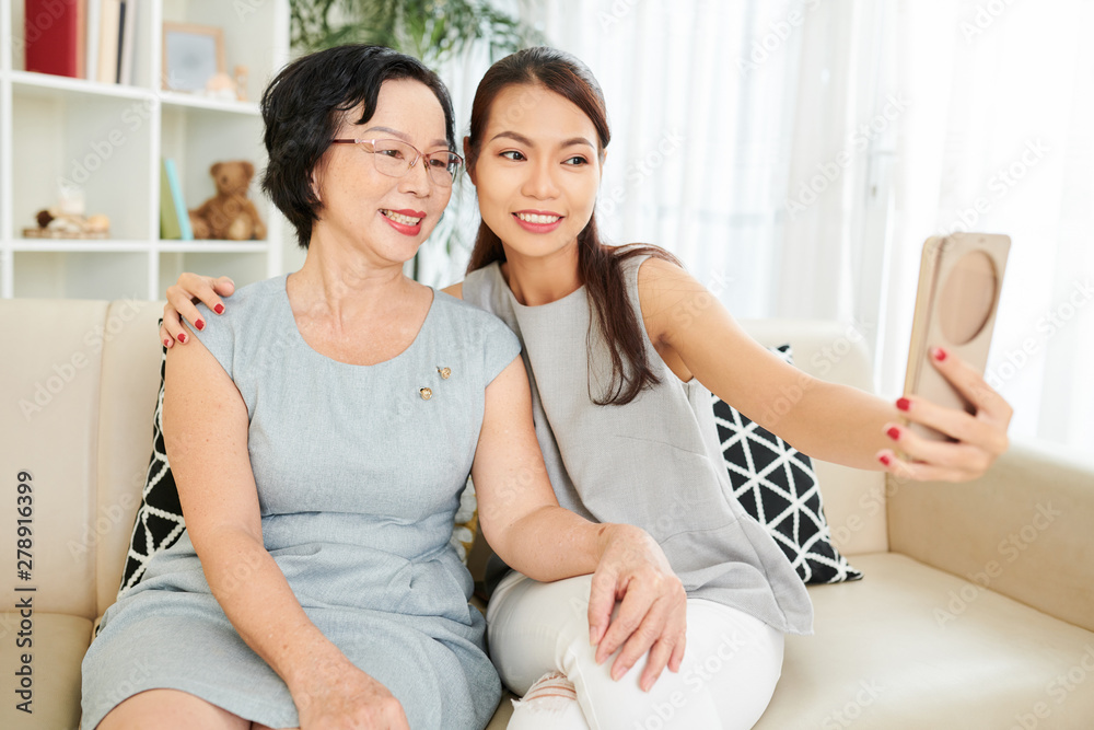 Young beautiful woman sitting on sofa embracing her mother in eyeglasses and making selfie portrait on her mobile phone Young beautiful woman sitting on sofa embracing her mother in eyeglasses and making selfie portrait on her mobile phone