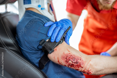 Medic wearing tourniquet on arm of injured person sitting on the driver saet after the road accident, providing emergency medical assistance