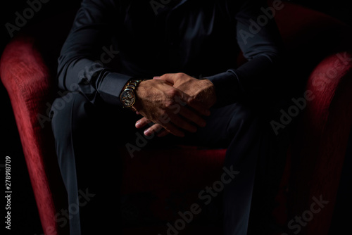 male businessman in a black suit sitting in red chair, black background, no faces visible, studio shooting