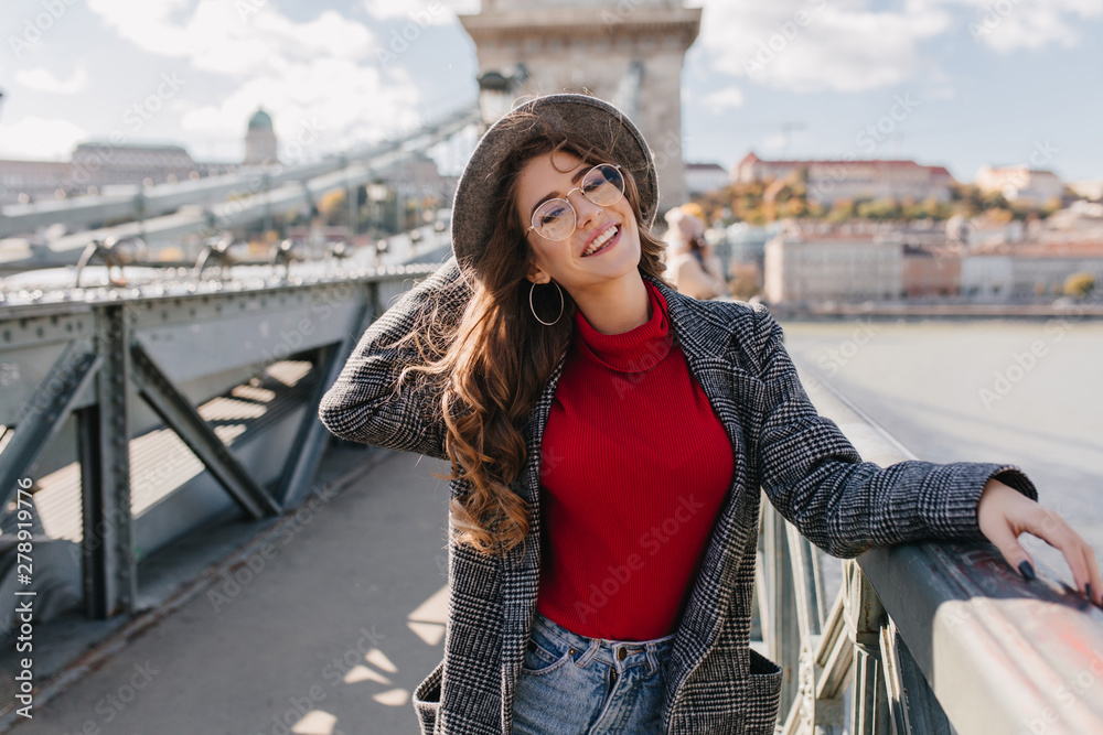 Fascinating girl in knitted red sweater playfully smiling on bridge in sunny day. Outdoor photo of dreamy brunette female model having fun in weekend in Europe.