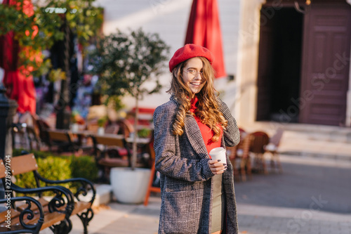 Stunning white girl in long coat drinking coffee while walking in park. Outdoor portrait of spectacular french woman with cup of latte standing on the street near cafe.