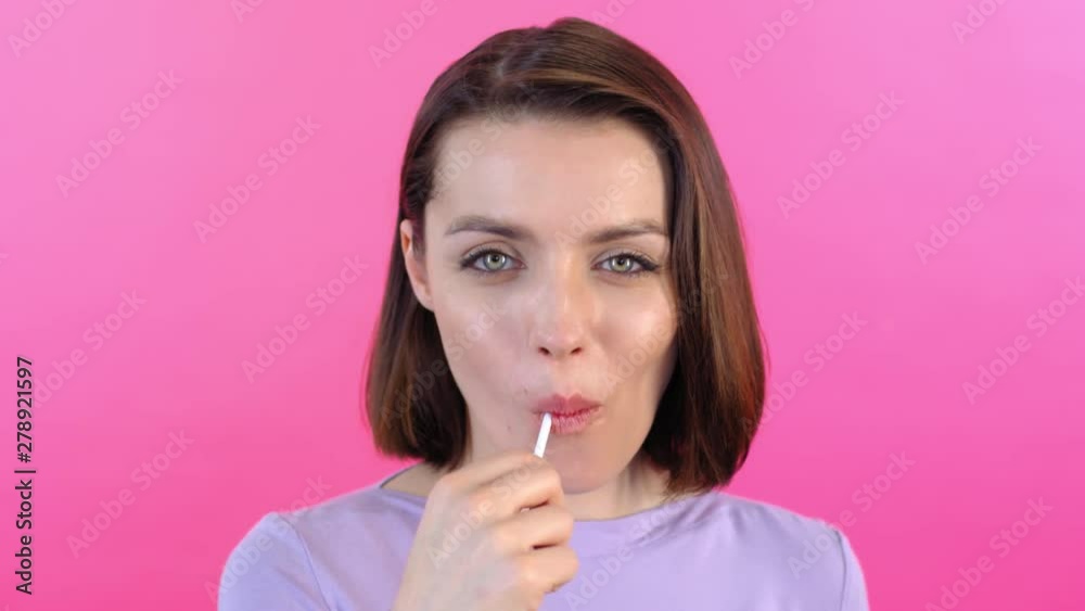 Close up face of attractive young woman enjoying sweet lollipop and smiling at camera on pink background Close up face of attractive young woman enjoying sweet lollipop and smiling at camera on pink background