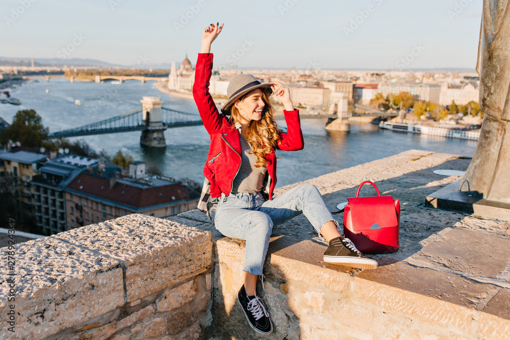 Adorable young female model with light-brown hair expressing happy emotions, traveling in Europe. Blissful girl in trendy red jacket and hat enjoying photoshoot on river background in city.