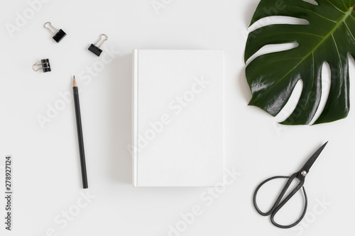 Top view of a white book mockup with  workspace accessories and a monstera leaf on a white table.