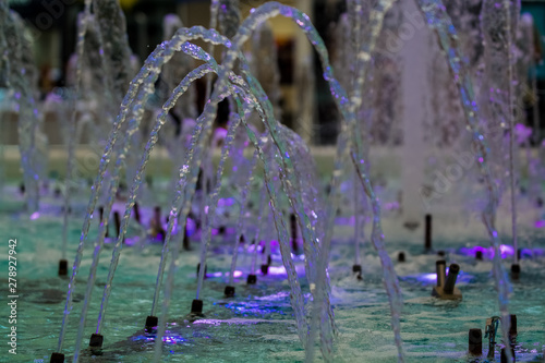 Big and strong water jets from the fountain with blue illumination in shopping center. Splashes and drops.