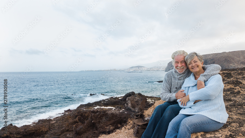 One senior couple of people waiting the sunrise sitting on the cliff and looking at camera. Early morning for two retired person in holiday at sea