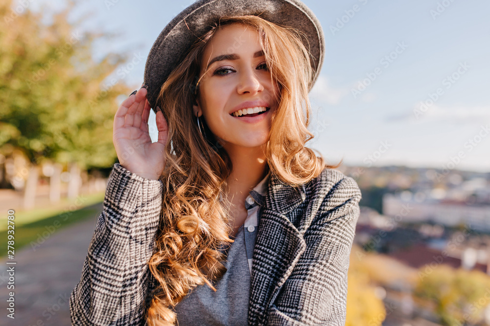 Magnificent young woman with beautiful blue eyes posing in hat on blur city background. Outdoor photo of ecstatic woman wears gray jacket walking around town in weekend.