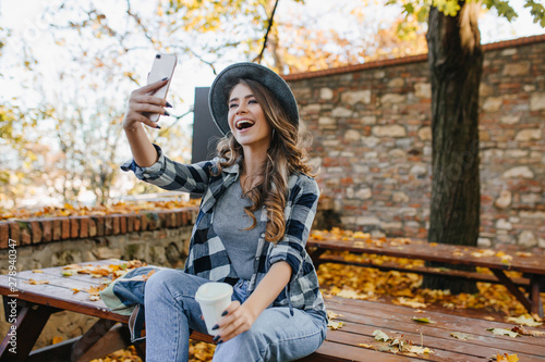 Positive woman with light-brown hair making selfie while drinking coffee in autumn park. Excited white lady in casual outfit relaxing in yard and taking picture of herself.