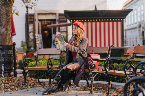 Worried girl in black leather boots looking at map sitting in park. Lost young woman in cute red beret trying to find her destination.