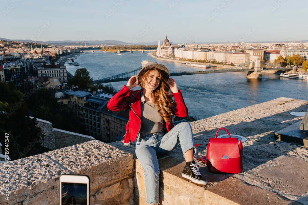 Blissful girl in casual clothes sitting on roof and touching her hat. Outdoor portrait of active brunette woman posing on beautiful river background.