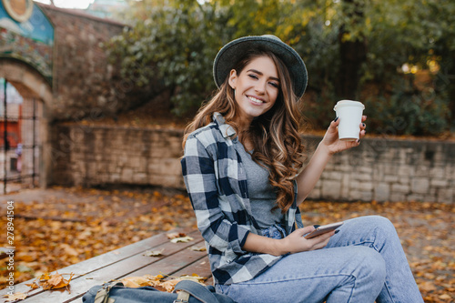 Playful girl in blue jeans sitting on wooden table and enjoying good autumn day. Pretty caucasian lady with light-brown hair drinks latte in yard in october morning.