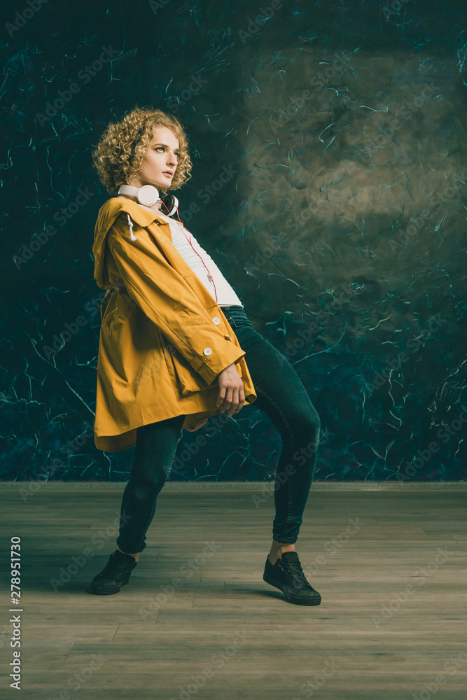 Obraz premium A young guy with a surfer's hair in a yellow jacket and headphones is dancing against a dark background