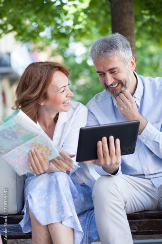 Happy mature tourists sitting on a bench in park and relaxing. Woman is holding a map while man is using a tablet