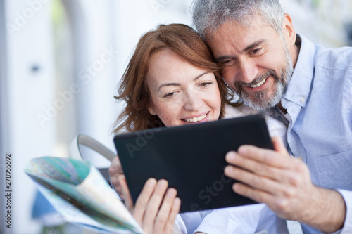 Loving mature couple sitting on a bench embraced in a hug, tenderly holding face to face and looking at photos on a tablet. Tourists using tablet