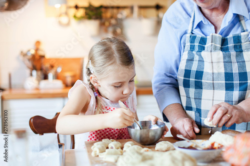 Family is cooking in cozy kitchen at home. Grandmother is teaching little girl. Retired woman and child make pastry dough together. Cute kid is helping to prepare meal. Lifestyle authentic moments.