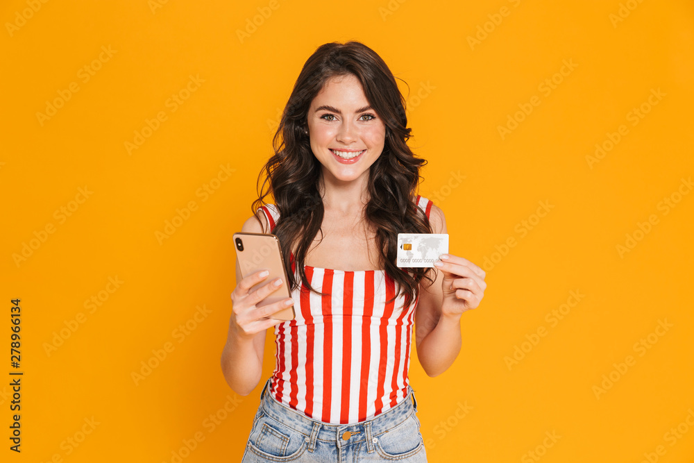 Happy young emotional girl isolated over yellow wall background using mobile phone holding credit card. Happy young emotional girl isolated over yellow wall background using mobile phone holding credit card.