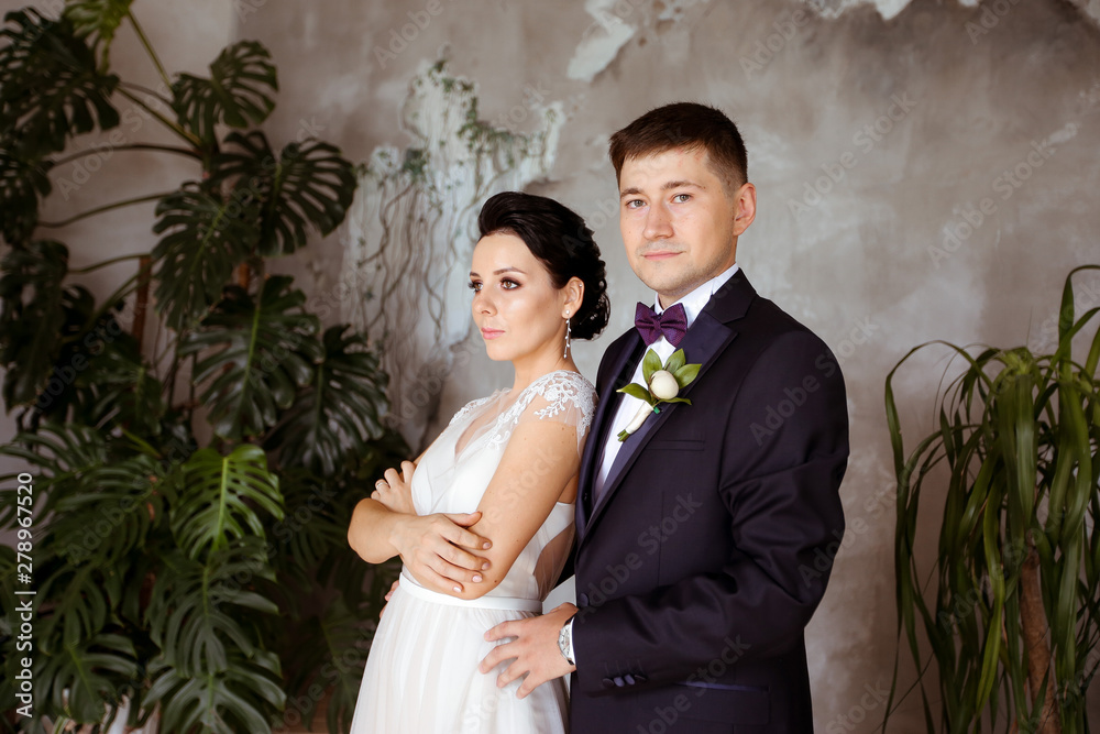 Bride in an elegant dress and groom in a suit on a background of tropical plants Bride in an elegant dress and groom in a suit on a background of tropical plants