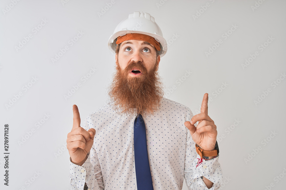 Young redhead irish architect man wearing security helmet over isolated white background amazed and surprised looking up and pointing with fingers and raised arms. Young redhead irish architect man wearing security helmet over isolated white background amazed and surprised looking up and pointing with fingers and raised arms.