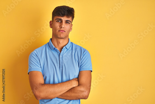 Young indian man wearing blue polo standing over isolated yellow background skeptic and nervous, disapproving expression on face with crossed arms. Negative person.