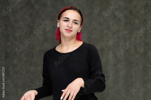 Portrait to the waist of a pretty girl with red hair on a gray background in a black jacket. Standing right in front of the camera in a studio with emotions, talking, showing hands, smiling