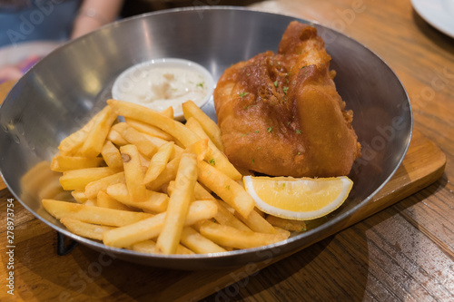 Fried fish steak served with french fries and tartar sauce in a deep pan placed on a wooden board.
