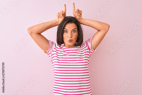 Young beautiful woman wearing striped t-shirt standing over isolated pink background doing funny gesture with finger over head as bull horns