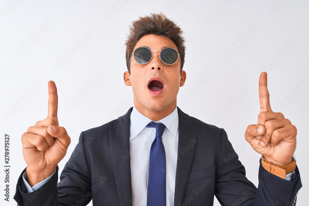 Young handsome businessman wearing suit and sunglasses over isolated white background amazed and surprised looking up and pointing with fingers and raised arms. Young handsome businessman wearing suit and sunglasses over isolated white background amazed and surprised looking up and pointing with fingers and raised arms.