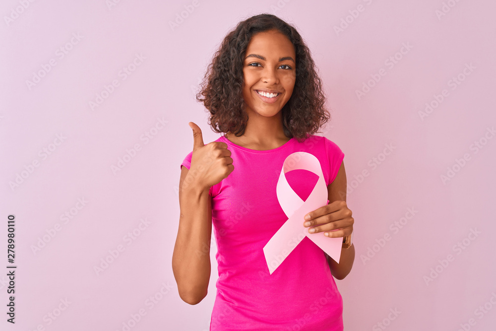 Young brazilian woman holding cancer ribbon standing over isolated pink background happy with big smile doing ok sign, thumb up with fingers, excellent sign