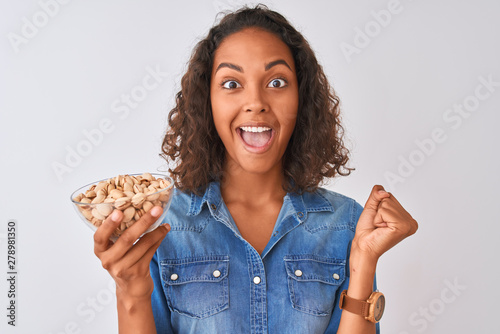Young brazilian woman holding bowl with pistachios standing over isolated white background screaming proud and celebrating victory and success very excited, cheering emotion