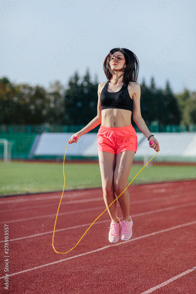 Sport, exercises outdoors. woman in black top and rose shorts jumping on skipping rope on stadium. Sporty girl in good shape, full body, outdoors