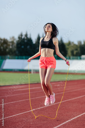 Achieving best results. Beautiful young brunette woman in sports clothing skipping rope and smiling while exercising on the running track outdoors © lena_itzy
