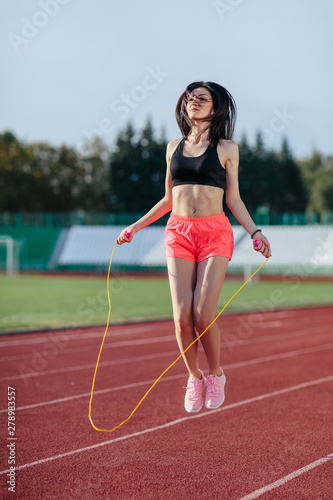 Sport, exercises outdoors. woman in black top and rose shorts jumping on skipping rope on stadium. Sporty girl in good shape, full body, outdoors © lena_itzy