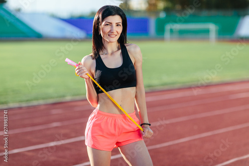 Portrait of happy young sports brunette woman in black top and rose shorts outdoors on stadium holding skipping rope. © lena_itzy