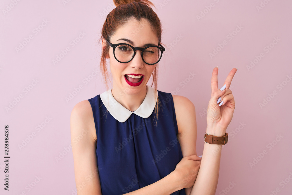 Redhead woman with pigtail wearing elegant dress and glasses over isolated pink background smiling with happy face winking at the camera doing victory sign. Number two. Redhead woman with pigtail wearing elegant dress and glasses over isolated pink background smiling with happy face winking at the camera doing victory sign. Number two.