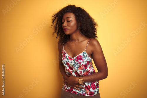African american woman wearing floral summer t-shirt over isolated yellow background with hand on stomach because nausea, painful disease feeling unwell. Ache concept.