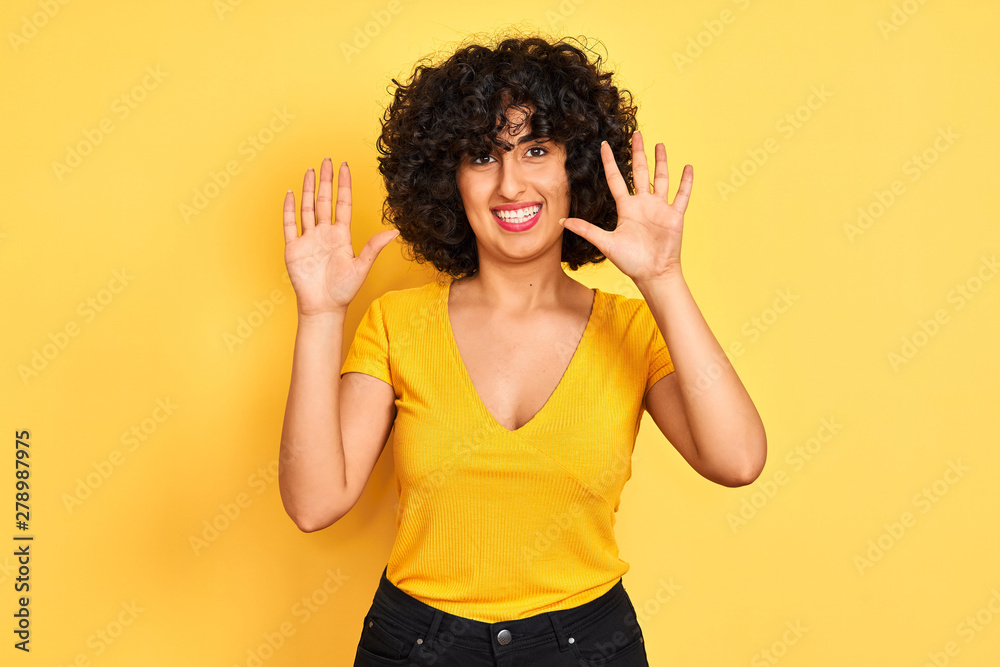 Young arab woman with curly hair wearing t-shirt standing over isolated yellow background showing and pointing up with fingers number ten while smiling confident and happy. Young arab woman with curly hair wearing t-shirt standing over isolated yellow background showing and pointing up with fingers number ten while smiling confident and happy.