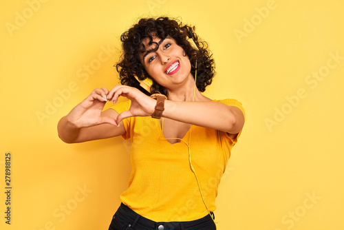 Arab woman with curly hair listening to music using headphones over isolated yellow background smiling in love showing heart symbol and shape with hands. Romantic concept.