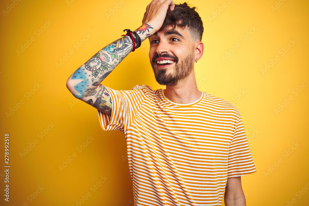 Young man with tattoo wearing striped t-shirt standing over isolated yellow background surprised with hand on head for mistake, remember error. Forgot, bad memory concept. Young man with tattoo wearing striped t-shirt standing over isolated yellow background surprised with hand on head for mistake, remember error. Forgot, bad memory concept.