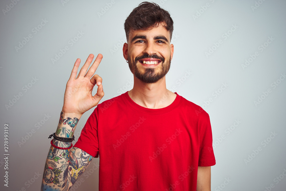Young man with tattoo wearing red t-shirt standing over isolated white background smiling positive doing ok sign with hand and fingers. Successful expression. Young man with tattoo wearing red t-shirt standing over isolated white background smiling positive doing ok sign with hand and fingers. Successful expression.