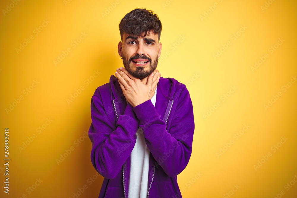 Young man with tattoo wearing sport purple sweatshirt over isolated yellow background shouting and suffocate because painful strangle. Health problem. Asphyxiate and suicide concept. Young man with tattoo wearing sport purple sweatshirt over isolated yellow background shouting and suffocate because painful strangle. Health problem. Asphyxiate and suicide concept.