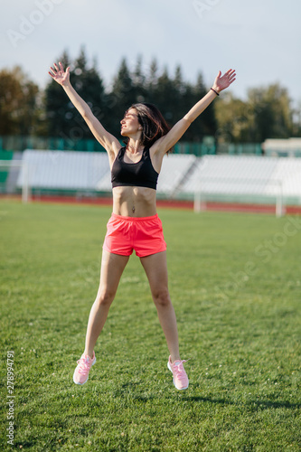 Attractive sporty brunette woman in pink shorts and top doing workout with jump rope in sun rays at the stadium