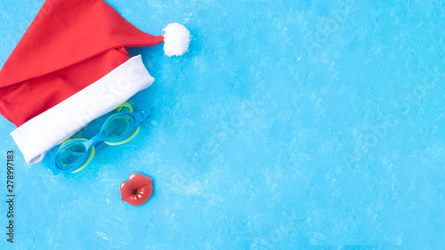 Banner. Hat of Santa Claus with goggles for swimming and a red lips. Christmas vacation, sandals and swimming glasses by water, slippers and pool goggles near swimming pool