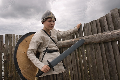 A teenager boy in a historical Viking costume with a helmet and a shield holding a sword standing in the middle of a wooden fortress at a historic festival.