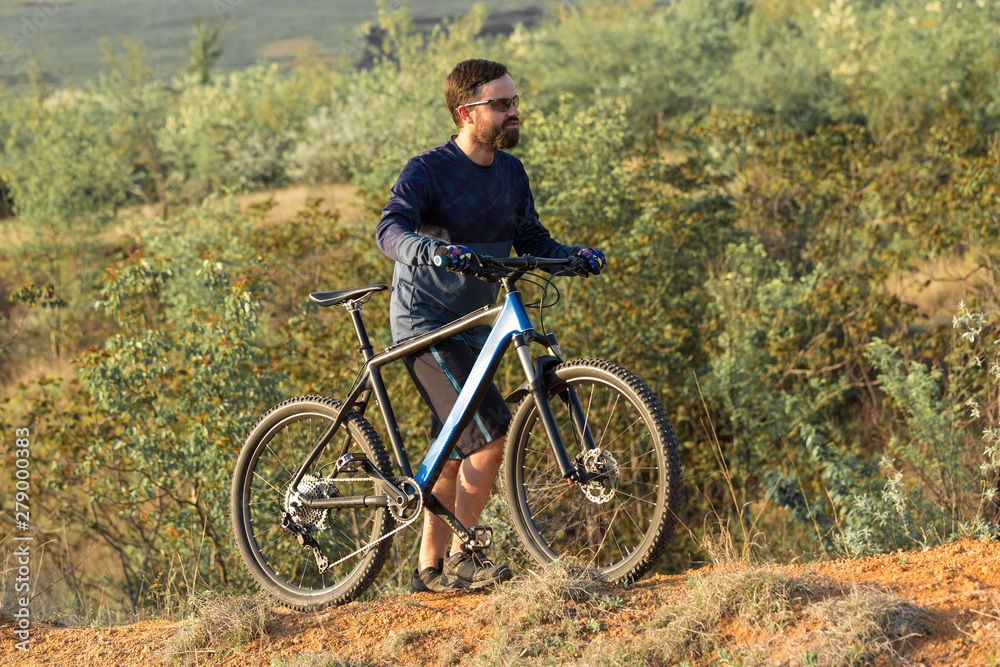 Cyclist in shorts and jersey on a modern carbon hardtail bike with an air suspension fork rides off-road on the orange-red hills at sunset evening in summer	