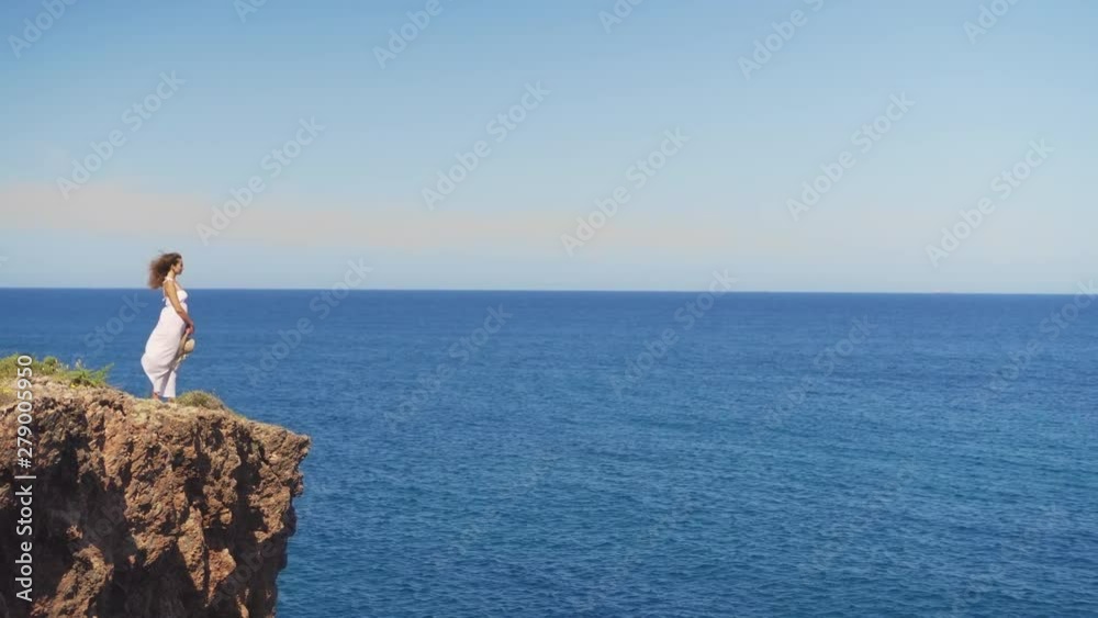 Girl in a white dress is standing on a rock cliff. a woman looks out into the distance on an endless sea surface Girl in a white dress is standing on a rock cliff. a woman looks out into the distance on an endless sea surface
