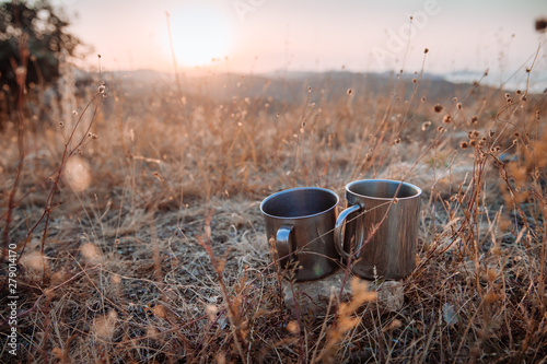 Two metal cups of tea among the grass against the backdrop of the mountains. Concept of autumn hiking trips