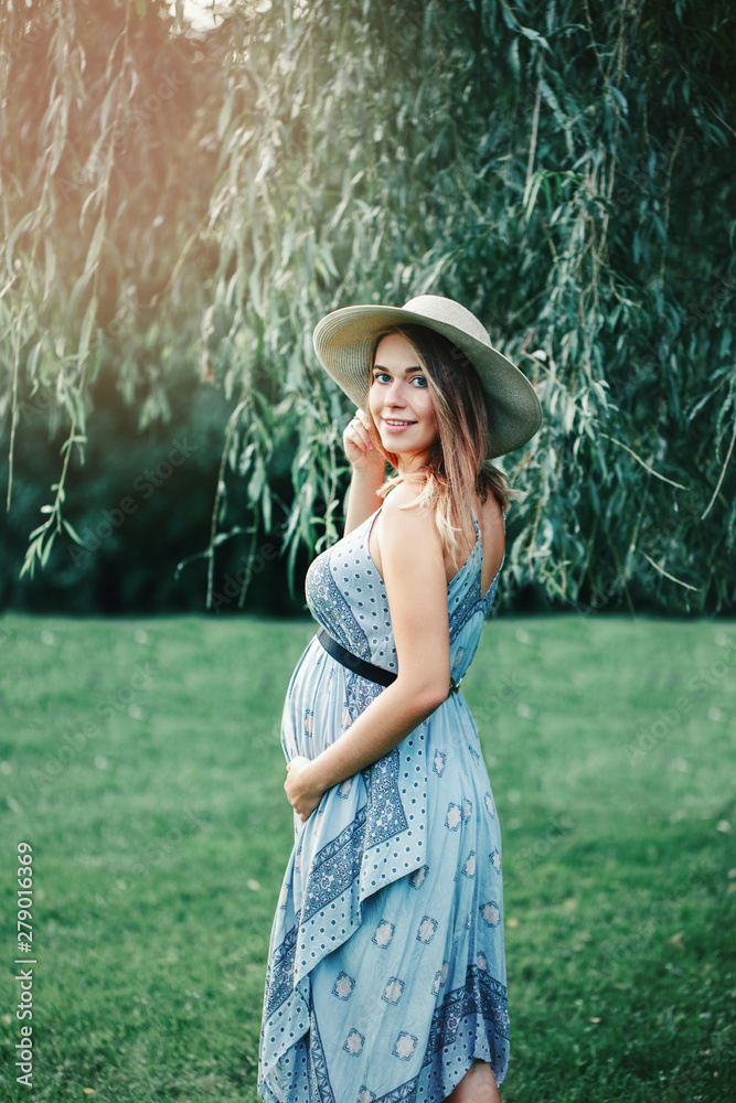 Foto de Happy healthy pregnancy and maternity. Portrait of pregnant young  Caucasian woman wearing long blue dress and rustic country hat in park  outside. Beautiful feminine expecting lady do Stock